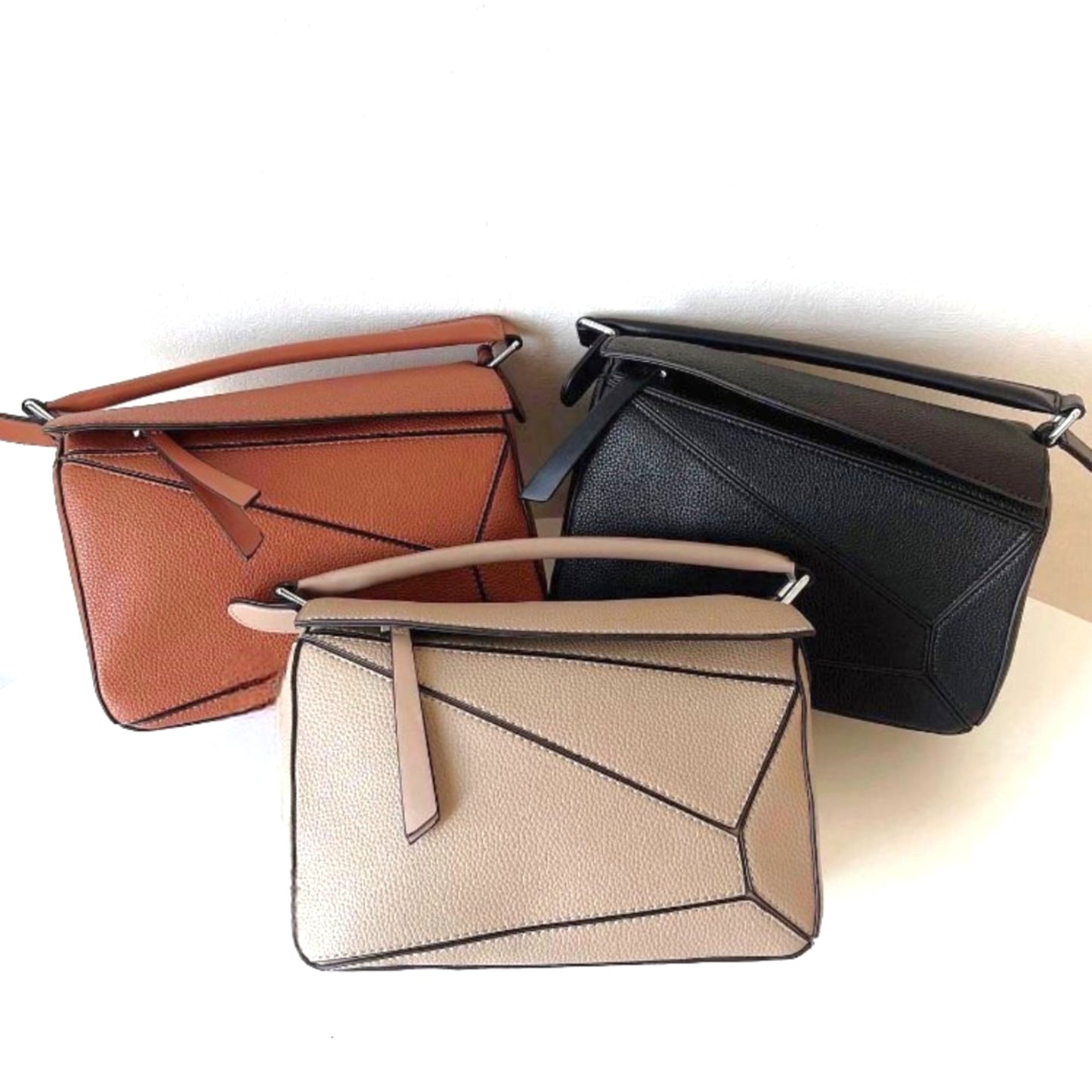 Amy, vegan leather handbags in Sand, Brown, and Black with geometric panels and top handles arranged in a group on display.