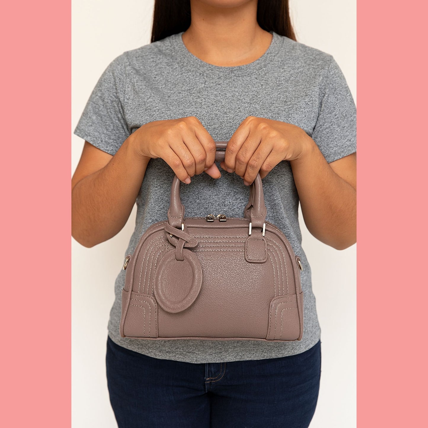 A model holding a modern and chic taupe Boston bowler bag by its top handles in front of her.