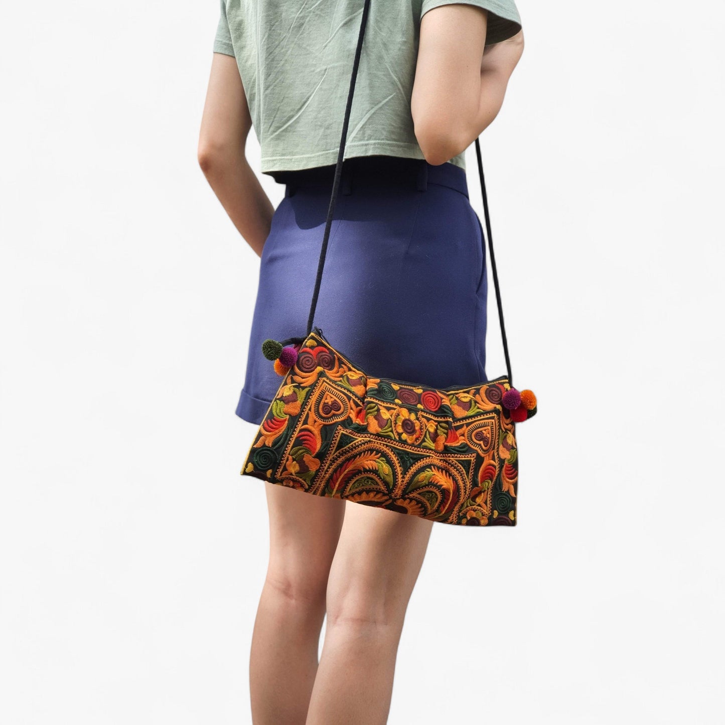 A woman carries an orange embroidered bag with colourful pom-poms and a skinny strap over her shoulder.