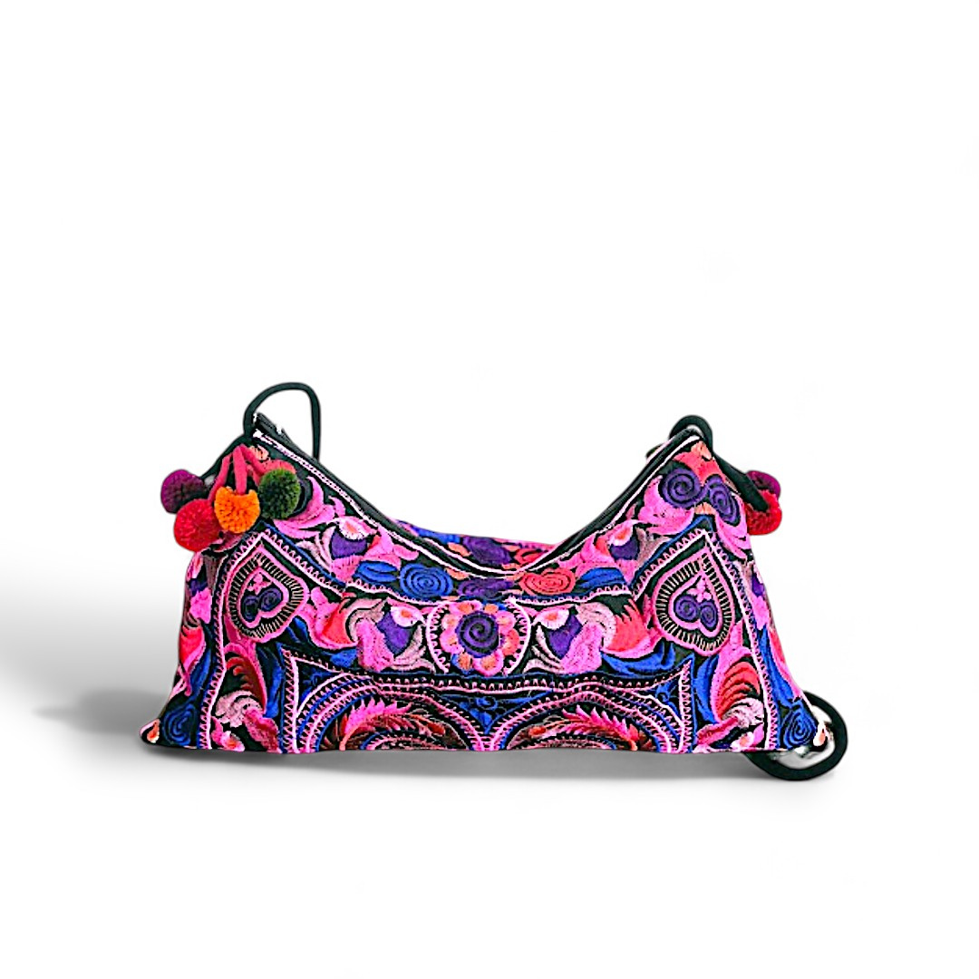 A pink crossbody bag with vibrant bird and floral embroidery, complemented by colourful pom-pom bag charms.