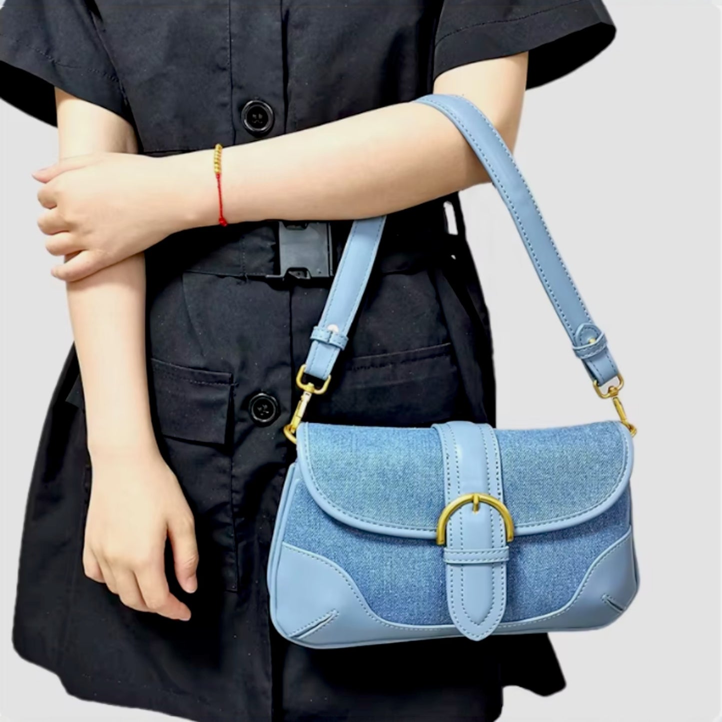 A woman carries a blue denim shoulder bag in the crook of her arm. The bag features leather trims and brass details.