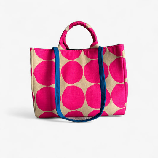 Front view of an oversized beige tote bag with big hot pink polka dots, top handles and contrasting blue straps.