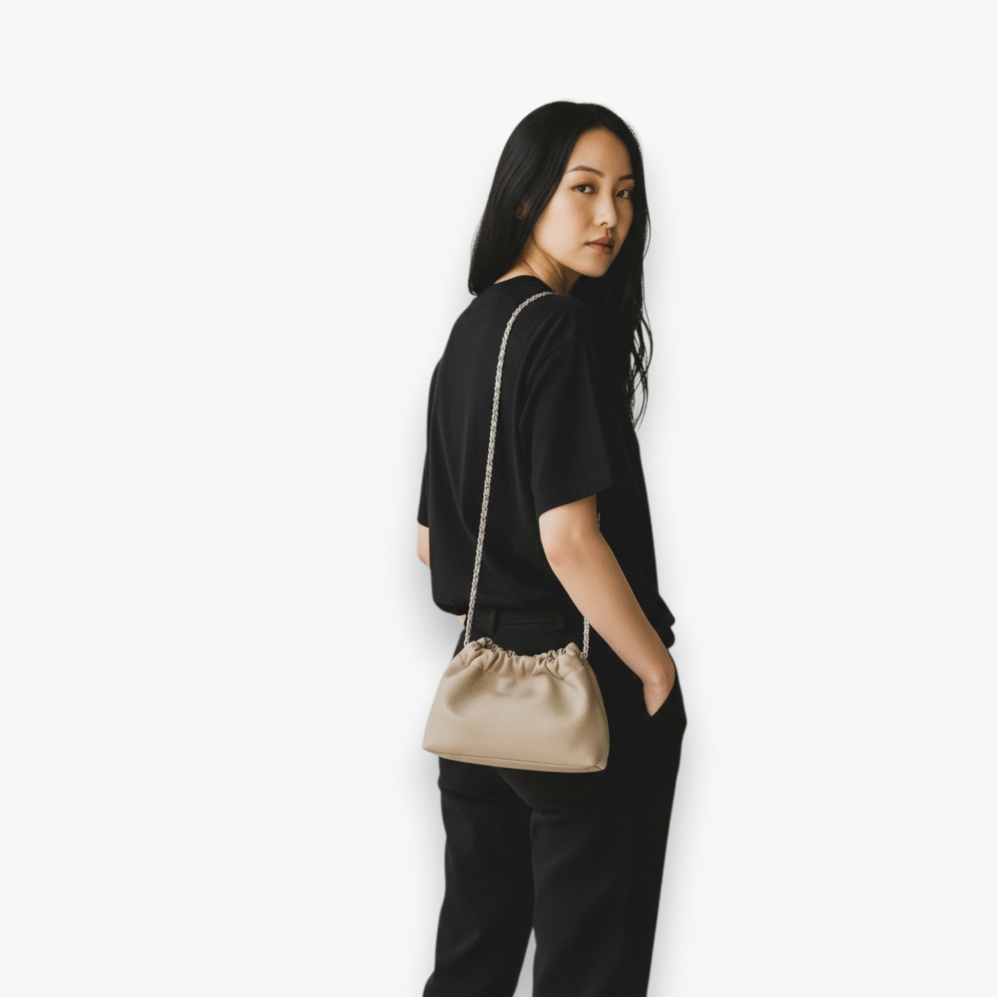 A woman carries a beige handbag with ruched-opening and a chain strap on her shoulder.