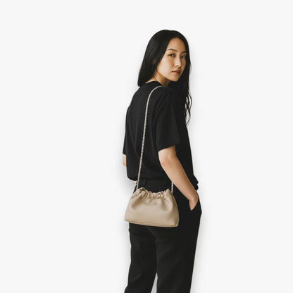 A woman carries a beige handbag with ruched-opening and a chain strap on her shoulder.