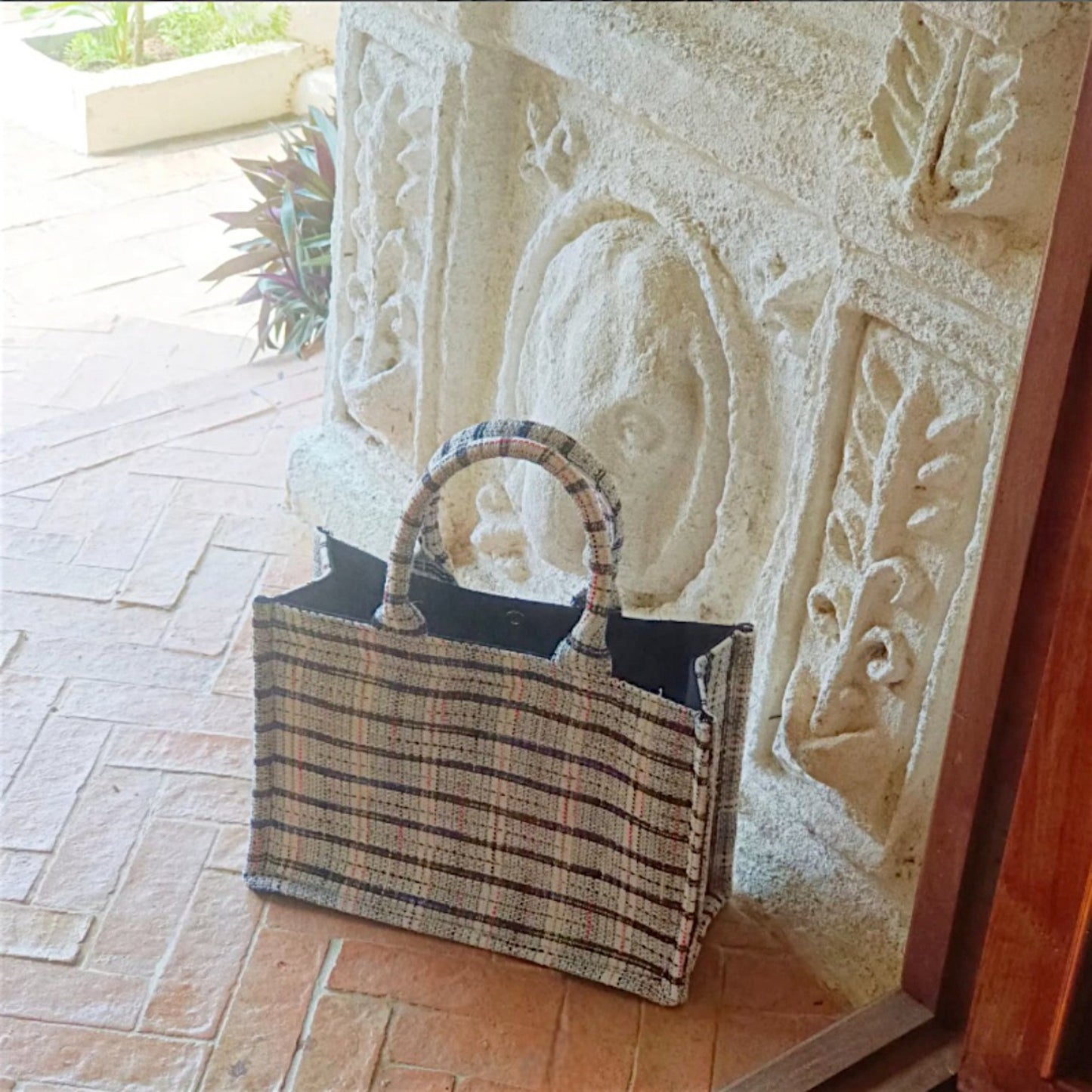 A sturdy tote with strong handles made with white tweed fabric with black, red, and blue threads is sitting outdoor.