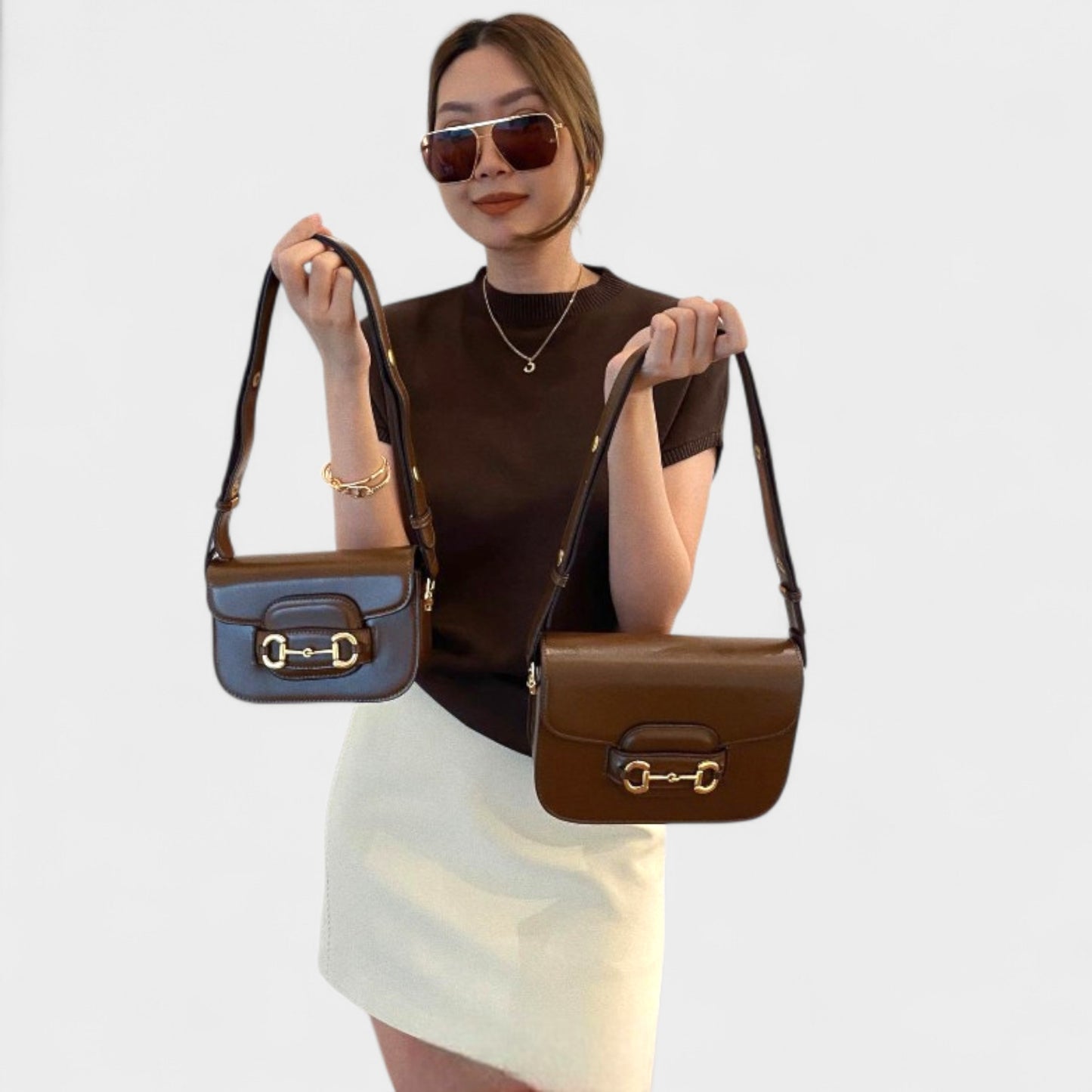 Model holds two brown equestrian-inspired leather shoulder bags, classic gold horse-bit buckle detail.